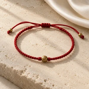Concept illustration: delicate red woven cord bracelet with a minimal antique gold tone bead and adjustable cord ends. Not an actual product photograph.